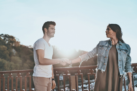 She is the one for him. Beautiful young couple holding hands and smiling while standing on the bridge outdoorsの写真素材