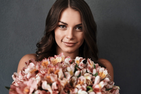 Real beauty. Attractive young woman with flower bouquet looking at camera and smiling while standing against grey backgroundの写真素材