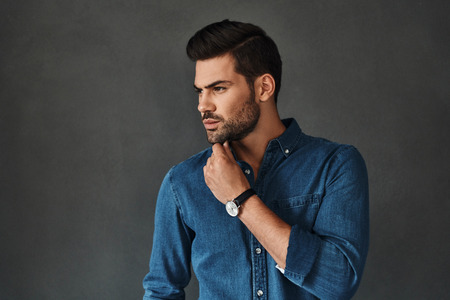 Thinking about... Serious young man keeping hand on chin and looking away while standing against grey backgroundの写真素材