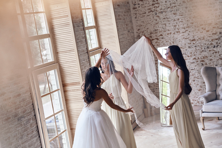 Her biggest day. Bridesmaids helping bride to put on a veil while standing near the window togetherの写真素材