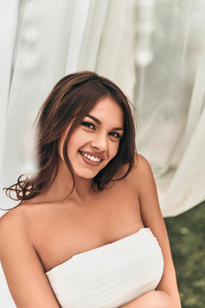 Attractive young woman in wedding dress looking at camera and smiling while standing outdoorsの写真素材