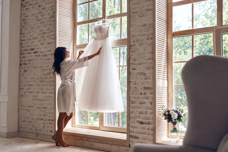 Full length of beautiful young woman in silk bathrobe touching her wedding dress while standing near the windowの写真素材