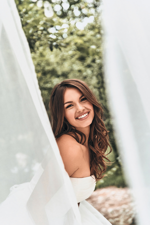 Attractive young woman in wedding dress looking at camera and smiling while standing outdoorsの写真素材