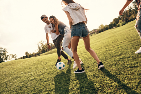 Trying to win. Group of young smiling people in casual wear enjoying nice summer day while playing soccer outdoorsの写真素材