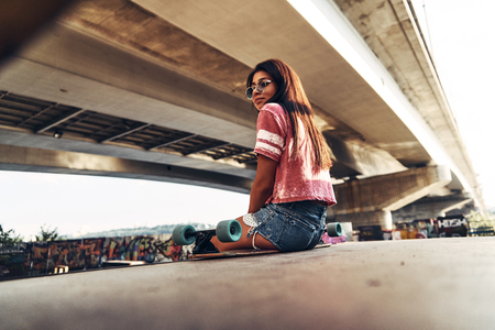 Time to relax. Attractive young woman sitting on the skateboard and looking away while spending time at the skate park outdoorsの写真素材