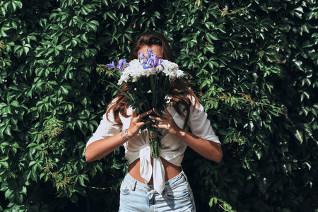 Just being herself. Close-up of attractive young woman in casual wear covering face with a bunch of flowers while standing in front of the ivy outdoorsの写真素材