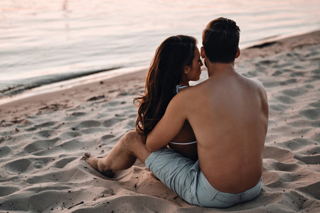 Two hearts full of love.  Rear view of young couple embracing while sitting on the river shoreの写真素材