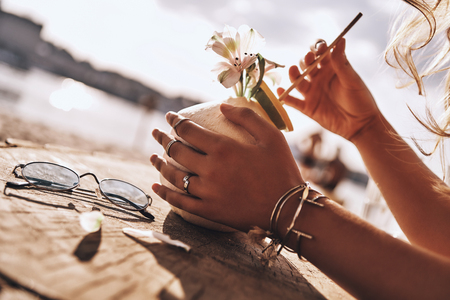 Tastes like summer! Close-up of young woman holding coconut cocktail with her hands while resting on the beachの写真素材
