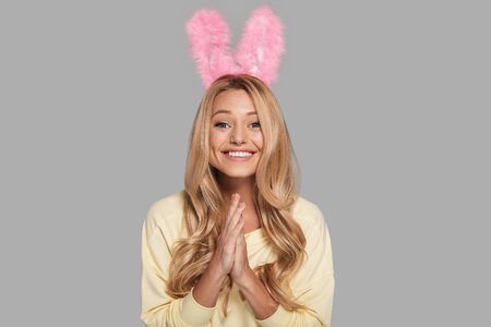 Feeling excited. Attractive young smiling woman in pink bunny ears keeping hands clasped and looking at camera while standing against grey backgroundの写真素材