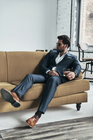Confident in his style. Good looking young man in full suit holding a glass and looking away while sitting on the sofaの写真素材