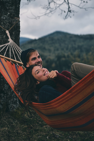 Totally happy. Beautiful young couple embracing and smiling while lying in hammock under the treeの写真素材