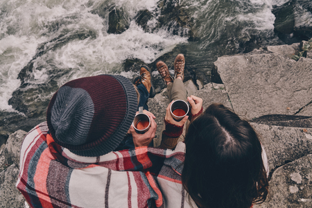 Warming up together. Close up top view of young couple covered with blanket holding cups while sitting on the rocks near the riverの写真素材