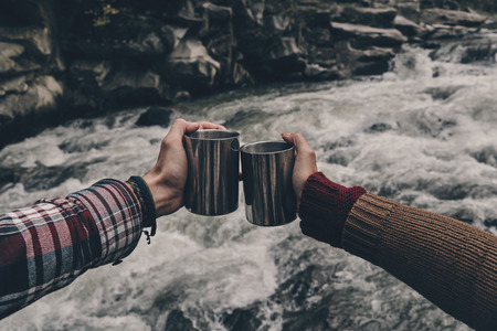 Cheers for love! Close up of young couple toasting each other while relaxing on the rocks near the riverの写真素材
