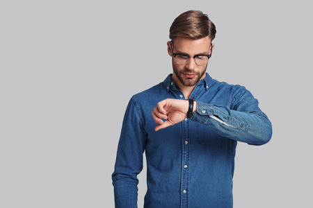 Time is money.  Thoughtful young man in eyewear checking the time while standing against grey backgroundの写真素材