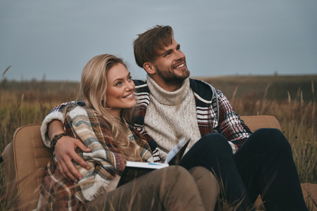 Enjoying their freedom.  Beautiful young couple embracing and looking away with smile while sitting on the fieldの写真素材