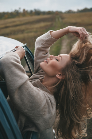 Feel the wind in your hair! Attractive young smiling woman leaning out the van?s window and keeping hand in hair while enjoying the car travelの写真素材