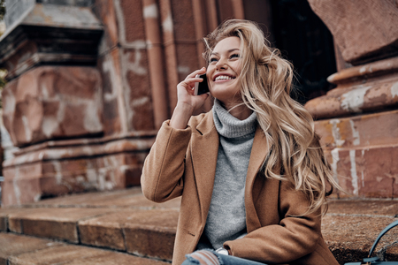 So good to hear you! Beautiful young woman talking on her smart phone and looking away with smile while sitting on stairs outdoorsの写真素材