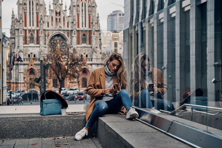 Searching for favorite song. Attractive young woman in coat listening music using her smart phone while spending carefree time in the cityの写真素材