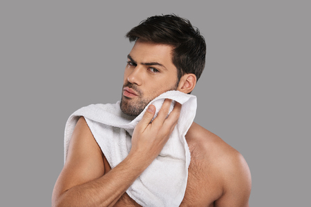 Everyday routine. Handsome young man wiping his face with towel while standing against grey backgroundの写真素材