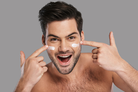 Amazing results! Handsome young smiling man applying moisturizer and looking at camera while standing against grey backgroundの写真素材