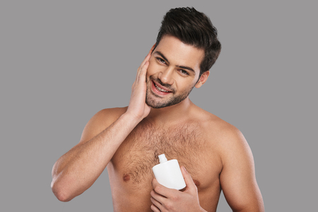Feeling so fresh. Handsome young man applying aftershave lotion and smiling while standing against grey backgroundの写真素材