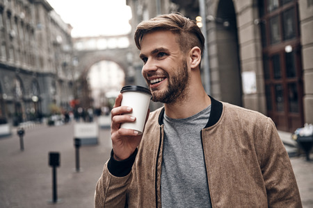 Enjoying fresh coffee. Handsome young man in casual wear holding disposable cup and smiling while walking through the city streetの写真素材