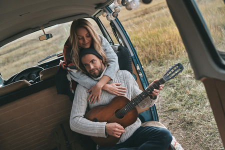 Playing romantic song.  Handsome young man playing guitar for his beautiful girlfriend while sitting in retro style mini vanの写真素材