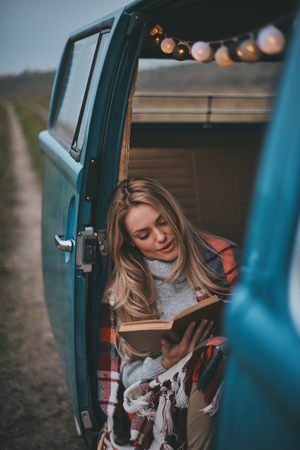 Involved in reading. Attractive young woman covered with blanket reading a book while sitting inside of the blue retro style mini vanの写真素材
