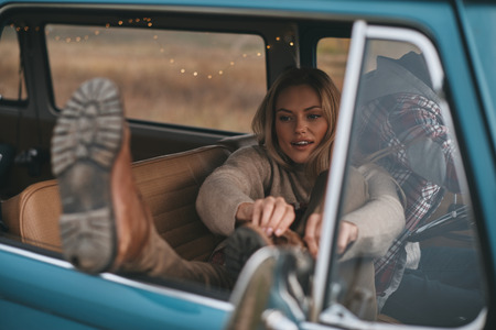 Carefree travel. Attractive young woman adjusting her boot while her boyfriend driving retro style mini vanの写真素材