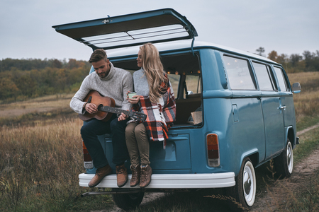 Romantic couple.  Handsome young man playing guitar for his beautiful girlfriend while sitting in the trunk of blue retro style mini vanの写真素材