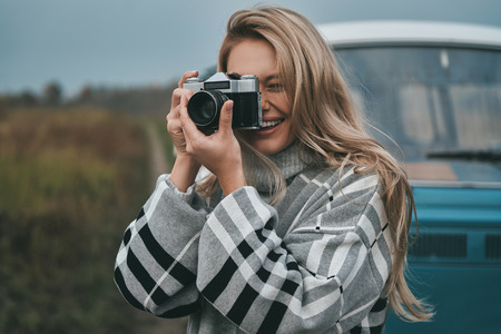 Cool shot! Attractive young woman photographing and smiling while standing outdoors with the blue retro style mini van in the backgroundの写真素材