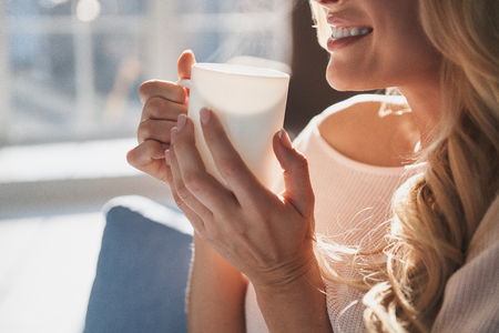 Enjoying morning coffee. Close up of young woman holding a cup and smiling while sitting on the sofa at homeの写真素材