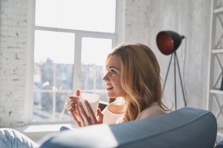 Quite contemplation. Attractive young woman holding a cup and looking away with smile while sitting...