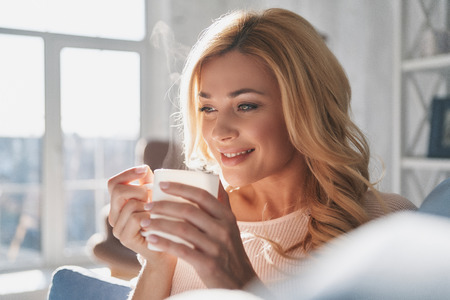 Dreaming about something. Attractive young woman holding a cup and looking away with smile while spending time at homeの写真素材