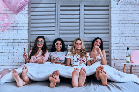 Friends forever. Four attractive young women in pajamas drinking cocktails and smiling while lying in the bedの写真素材