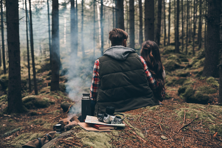 Rear view of young couple warming up by the campfire while sitting in the forestの写真素材