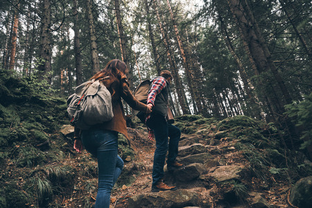 Rear view of modern young couple holding hands and moving up while hiking together in the woodsの写真素材