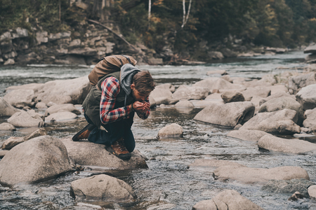 Handsome young modern man drinking water from the river while hiking in mountainsの写真素材