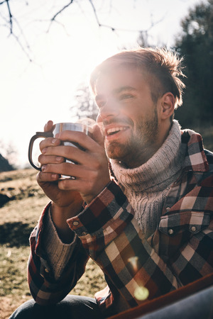 Handsome young smiling man in warm clothing having morning coffee while camping in mountainsの写真素材