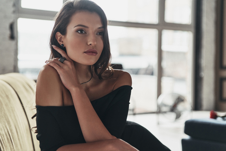 Gorgeous beauty. Attractive young woman in elegant black dress keeping hand in hair and looking away while sitting on the sofaの写真素材