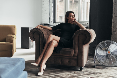 Quite contemplation. Attractive young woman in elegant black dress keeping hand on chin and looking away while sitting in the armchairの写真素材