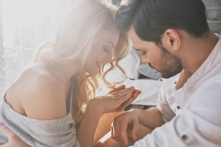 Pure feelings. Beautiful young woman looking at her engagement ring and smiling while sitting in the bedroom with her boyfriendの写真素材
