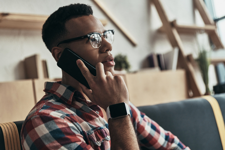 Listening to clients requirements. Thoughtful young man talking on his smart phone and looking away while sitting in the creative working spaceの写真素材