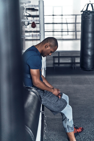 Making a pause. Tired young African man in sport clothing resting while sitting in the gymの写真素材