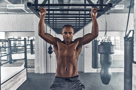Determined to win. Shirtless young African man looking at camera and doing pull-ups while exercising...