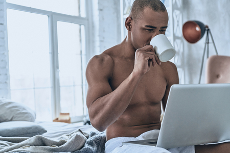 Searching for fresh news online. Handsome young African man drinking coffee and using computer while sitting on bed at homeの写真素材
