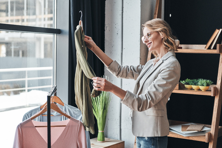 Young perfectionist. Attractive young woman in smart casual wear choosing clothes from the rank and smiling while standing in the workshopの写真素材