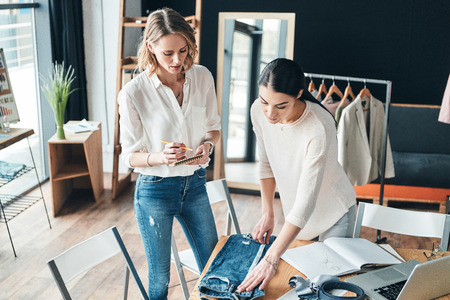 Working on new order. Top view of beautiful young women measuring jeans with tape while spending time in the workshopの写真素材