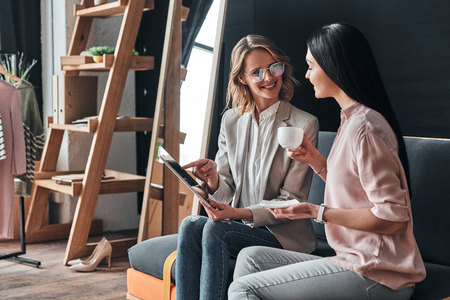 Discussing details with a client. Beautiful young woman showing something to her customer using digital tablet while sitting in the fashion workshopの写真素材