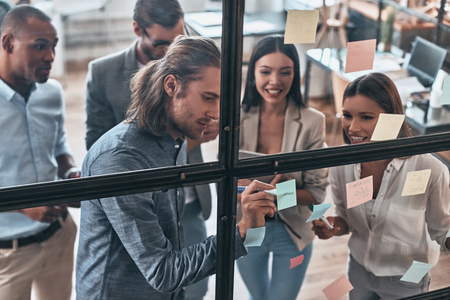 Happy business partners. Top view of young modern people in smart casual wear using adhesive notes while standing behind the glass wall in the board roomの写真素材